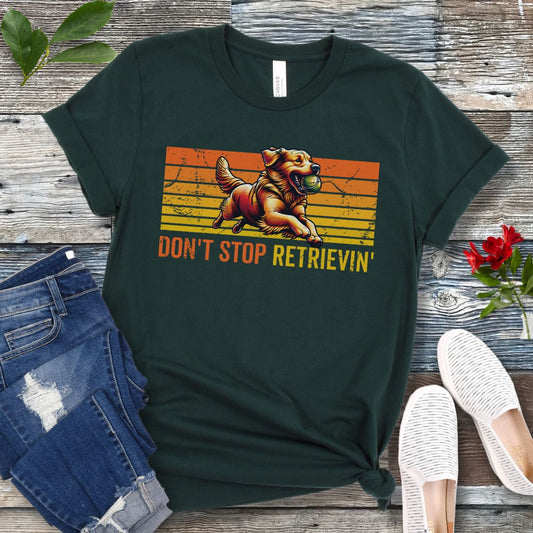 Dark green t-shirt with vintage sunset style horizontal stripes, a Golden Retriever running with a tennis ball, and the humorous text 'Don't Stop Retrieving', shown on a wooden surface with jeans and shoes.