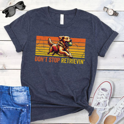 Heather navy t-shirt with vintage sunset style horizontal stripes, a Golden Retriever running with a tennis ball, and the humorous text 'Don't Stop Retrieving', shown on a wooden surface with jeans and shoes.