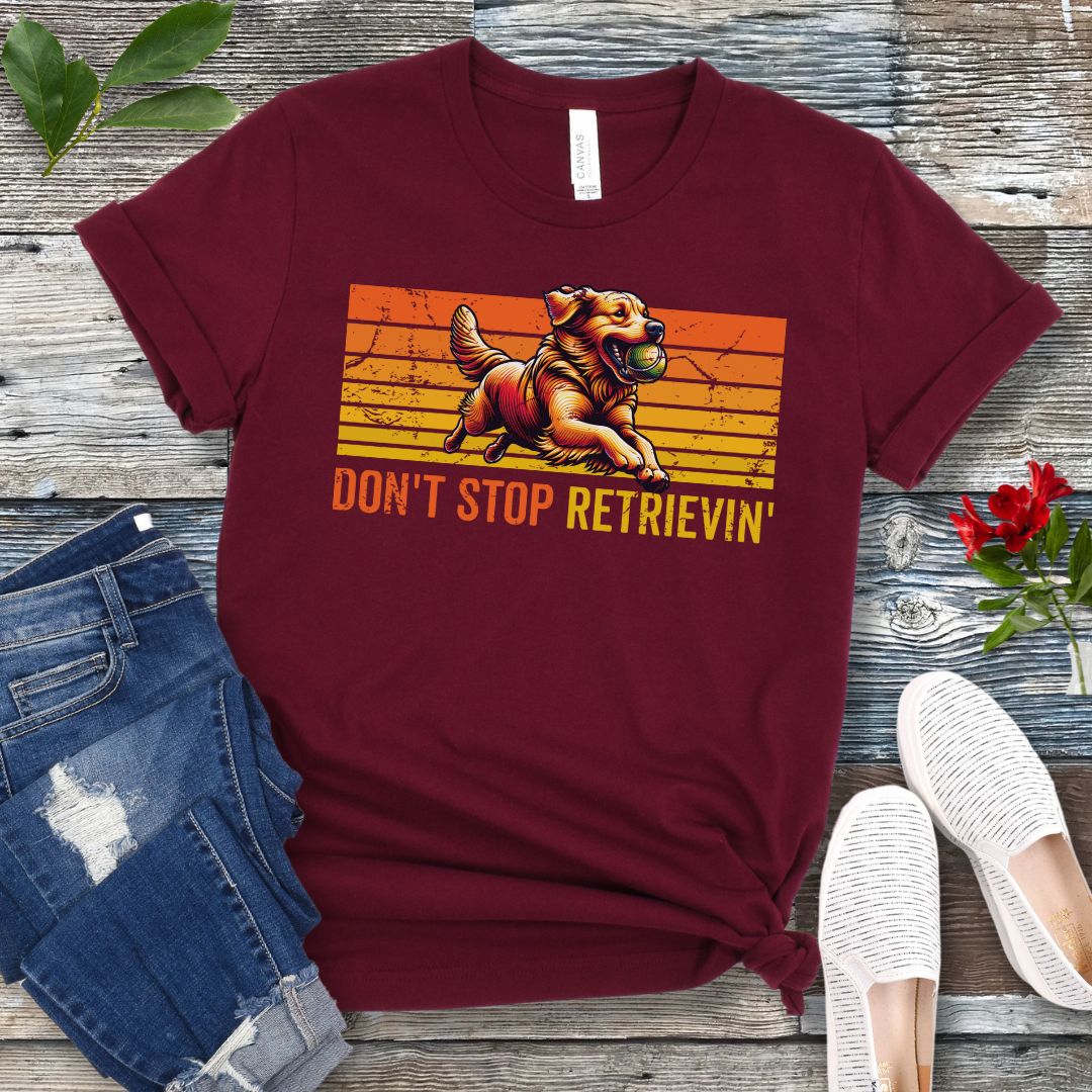 Maroon t-shirt with vintage sunset style horizontal stripes, a Golden Retriever running with a tennis ball, and the humorous text 'Don't Stop Retrieving', shown on a wooden surface with jeans and shoes.