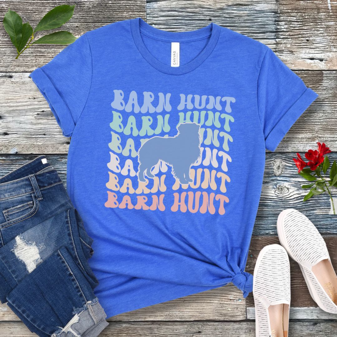 Royal blue dog sports t-shirt with 'Barn Hunt' repeated in colorful retro style text and a custom dog breed silhouette, presented on a wooden surface with jeans and shoes.