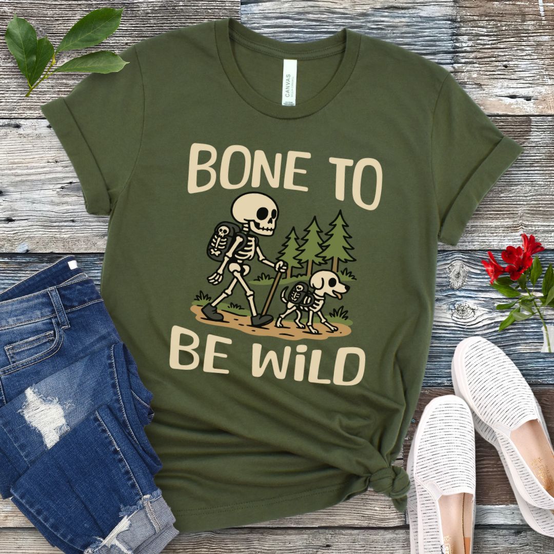 A green-colored t-shirt with a graphic print of a skeleton and a dog on a trail, with text reading 'BONE TO BE WILD'. The t-shirt is presented flat with a pair of blue jeans and white shoes in the background.