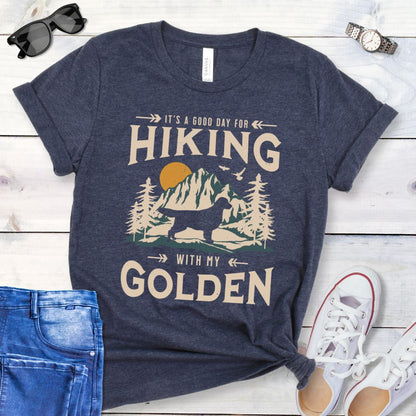 Heather navy Golden Retriever t-shirt with 'Hiking with my Golden' text, mountain scene and Golden Retriever silhouette, presented on a wooden surface with jeans and shoes.