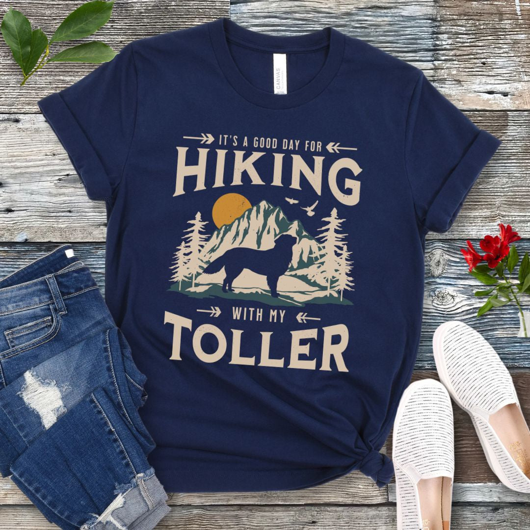 A navy blue t-shirt with a vintage-style graphic print of mountains and a Nova Scotia Duck Tolling Retriever and the text 'Hiking with my Toller'. The shirt is presented on a wooden surface with a pair of blue jeans and white shoes in the background.