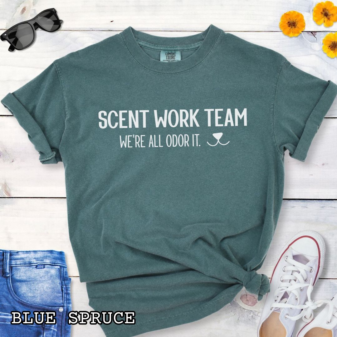 Green Comfort Colors t-shirt for dog scent trainers with the humorous text 'Scent Work Team, We're All Odor It' in bold lettering, shown on a wooden surface with sunglasses, jeans, and sneakers.