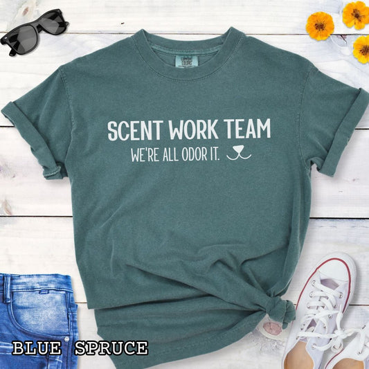 Green Comfort Colors t-shirt for dog scent trainers with the humorous text 'Scent Work Team, We're All Odor It' in bold lettering, shown on a wooden surface with sunglasses, jeans, and sneakers.