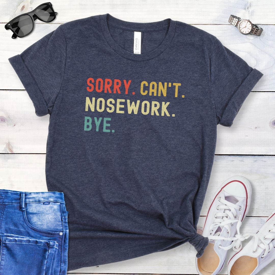Heather navy dog sports t-shirt with colorful retro text 'SORRY. CAN'T. NOSEWORK. BYE', shown on a wooden surface with jeans and shoes.