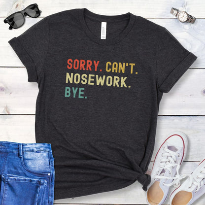 Dark grey dog sports t-shirt with colorful retro text 'SORRY. CAN'T. NOSEWORK. BYE', shown on a wooden surface with jeans and shoes.