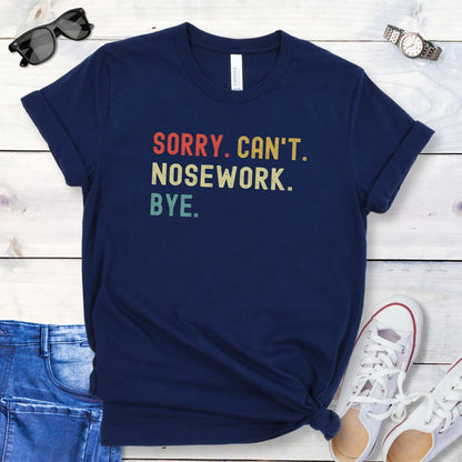 Navy dog sports t-shirt with colorful retro text 'SORRY. CAN'T. NOSEWORK. BYE', shown on a wooden surface with jeans and shoes.