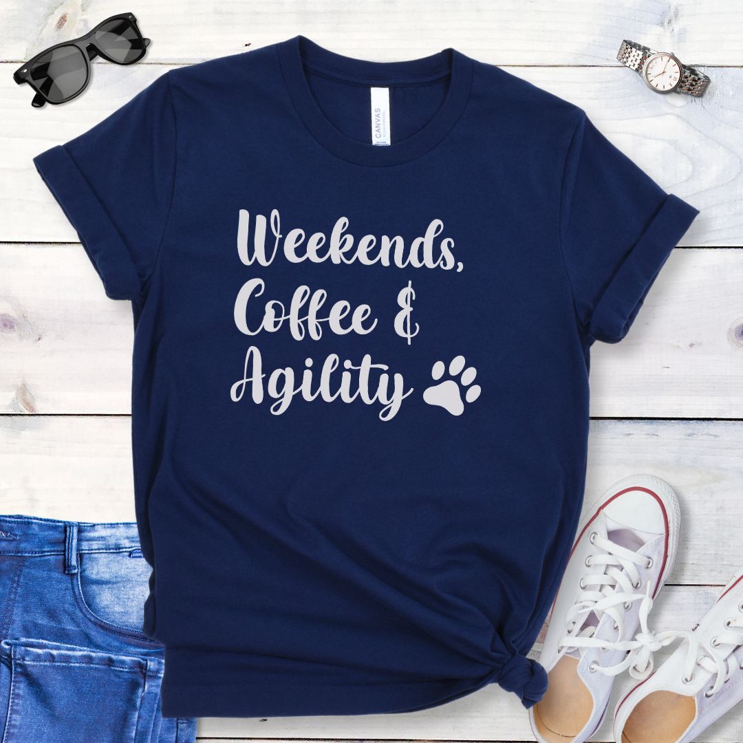 Navy dog agility t-shirt with 'Weekends, Coffee & Agility' text in playful script lettering, accented by a paw print, shown on a wooden surface with casual items.