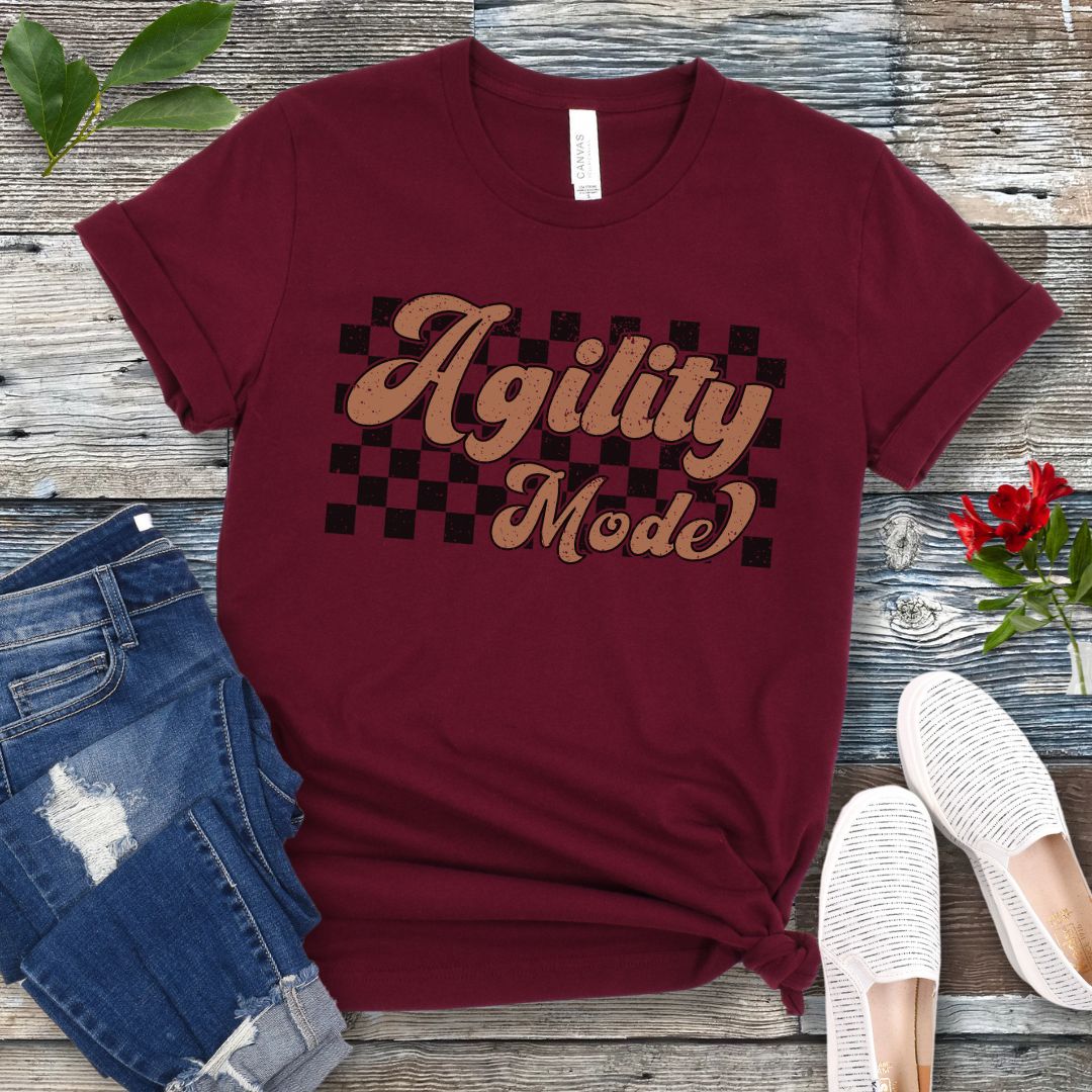 A maroon dog agility t-shirt with 'Agility Mode' written in a retro stylized font over a checkerboard pattern, placed on a wooden surface, accompanied by jeans and white shoes.