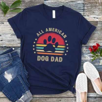 A navy unisex short sleeve T-shirt with 'ALL AMERICAN DOG DAD' printed on the front, accompanied by a colorful retro sunset graphic, a paw print and stars. The shirt is displayed flat with a pair of blue jeans and a pair of white shoes.