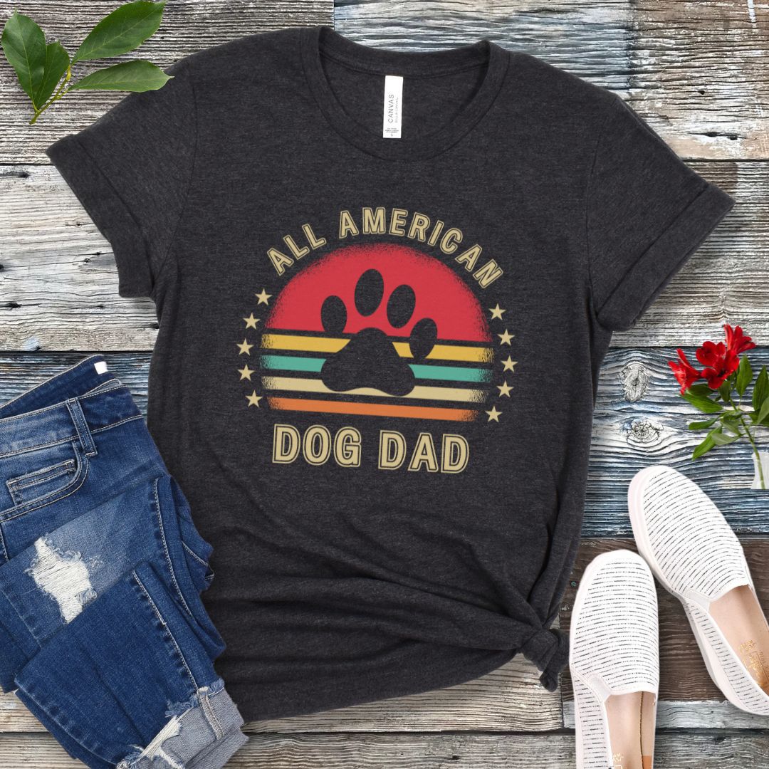 A dark grey unisex short sleeve T-shirt with 'ALL AMERICAN DOG DAD' printed on the front, accompanied by a colorful retro sunset graphic, a paw print and stars. The shirt is displayed flat with a pair of blue jeans and a pair of white shoes.