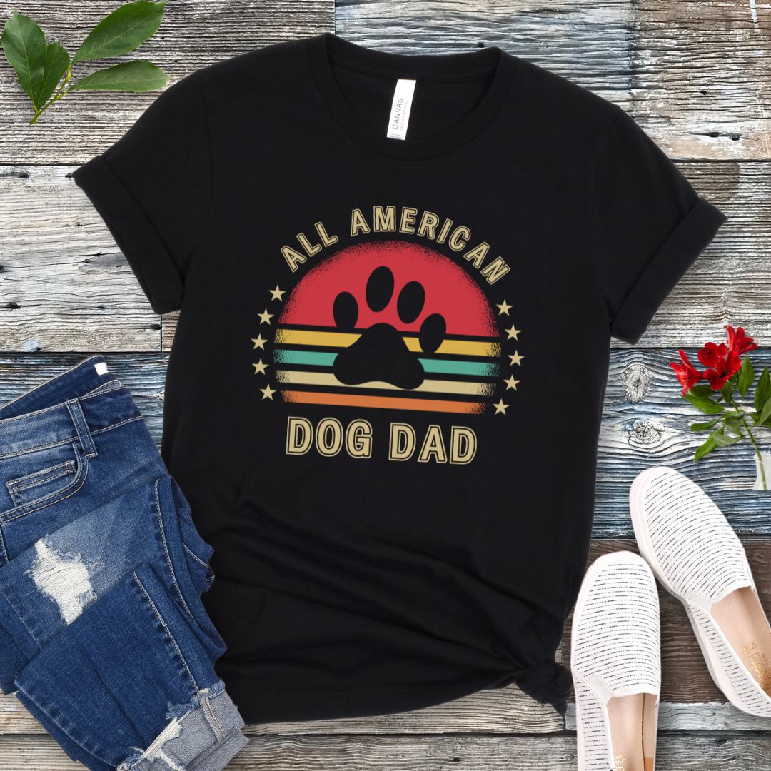 A black unisex short sleeve T-shirt with 'ALL AMERICAN DOG DAD' printed on the front, accompanied by a colorful retro sunset graphic, a paw print and stars. The shirt is displayed flat with a pair of blue jeans and a pair of white shoes.