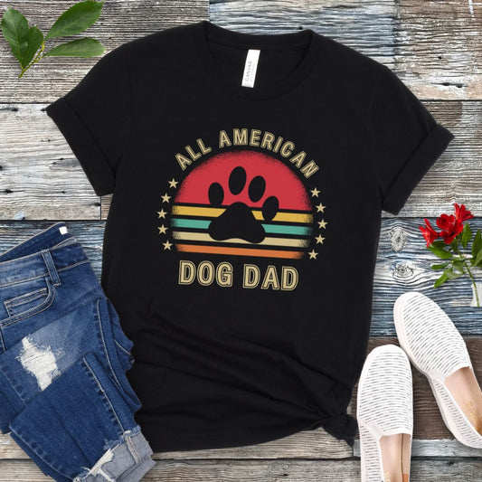 A black unisex short sleeve T-shirt with 'ALL AMERICAN DOG DAD' printed on the front, accompanied by a colorful retro sunset graphic, a paw print and stars. The shirt is displayed flat with a pair of blue jeans and a pair of white shoes.