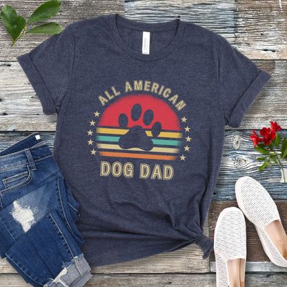 A heather navy unisex short sleeve T-shirt with 'ALL AMERICAN DOG DAD' printed on the front, accompanied by a colorful retro sunset graphic, a paw print and stars. The shirt is displayed flat with a pair of blue jeans and a pair of white shoes.