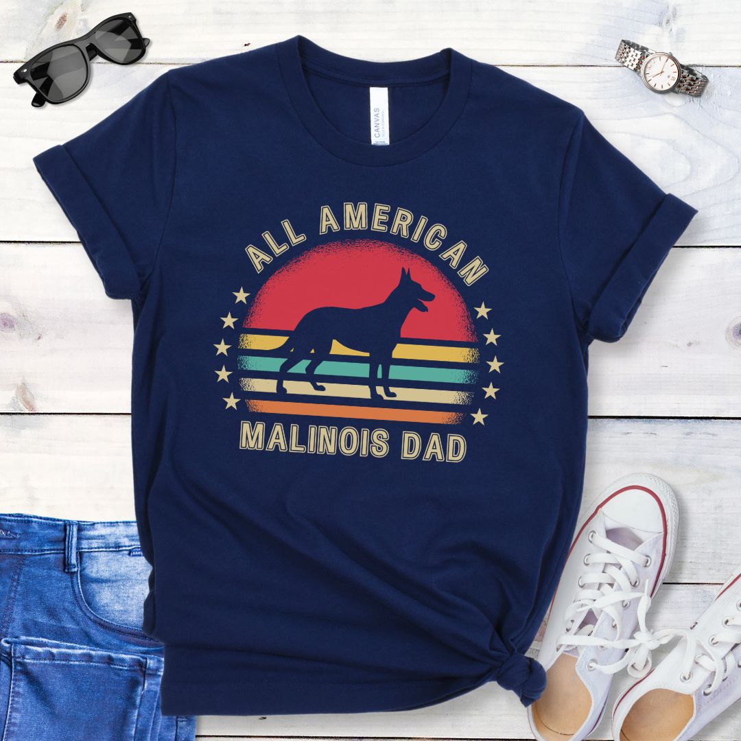 A navy blue dog lover t-shirt with 'All American Malinois Dad' printed on it, featuring a silhouette of a Belgian Malinois dog, a colorful vintage sunset, and stars. The shirt is presented on a wooden surface with denim jeans and white shoes in the background.