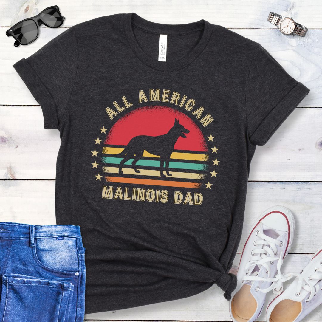 A dark grey dog lover t-shirt with 'All American Malinois Dad' printed on it, featuring a silhouette of a Belgian Malinois dog, a colorful vintage sunset, and stars. The shirt is presented on a wooden surface with denim jeans and white shoes in the background.
