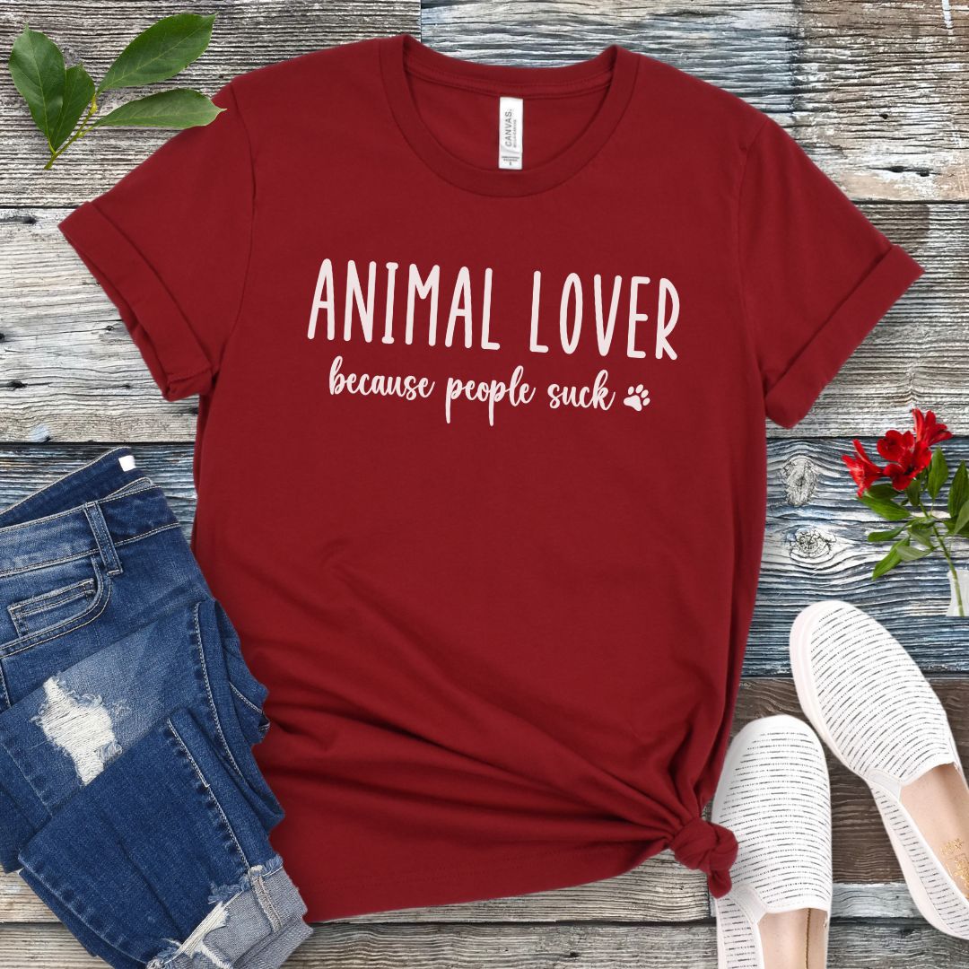 Dark red unisex t-shirt with 'Animal Lover because people suck' text in playful lettering, shown on a wooden surface with casual items.