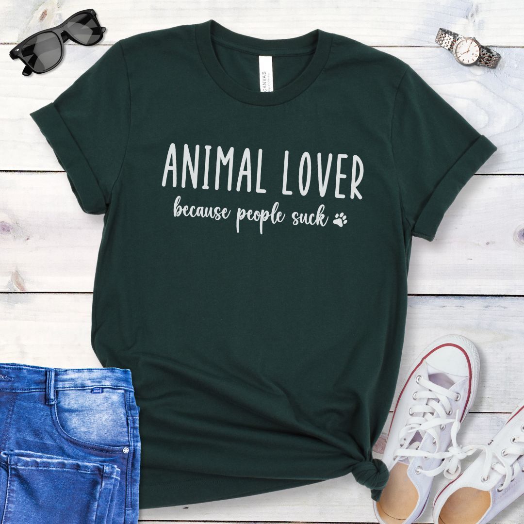 Dark green unisex t-shirt with 'Animal Lover because people suck' text in playful lettering, shown on a wooden surface with casual items.