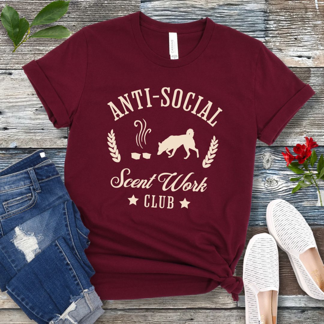 Maroon K9 nosework t-shirt with 'Anti-Social Scent Work Club' text and graphic of dog sniffing scentwork hides, on a wooden surface with jeans and shoes.