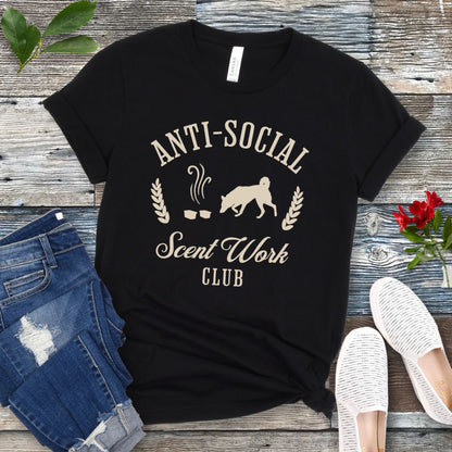 Black K9 nosework t-shirt with 'Anti-Social Scent Work Club' text and graphic of dog sniffing scentwork hides, on a wooden surface with jeans and shoes.