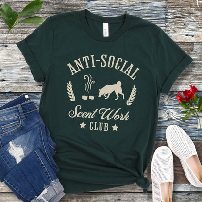 Dark green K9 nosework t-shirt with 'Anti-Social Scent Work Club' text and graphic of dog sniffing scentwork hides, on a wooden surface with jeans and shoes.