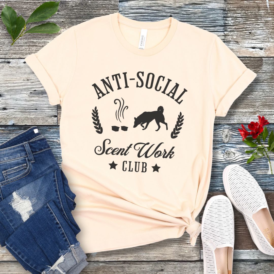 Beige K9 nosework t-shirt with 'Anti-Social Scent Work Club' text and graphic of dog sniffing scentwork hides, on a wooden surface with jeans and shoes.