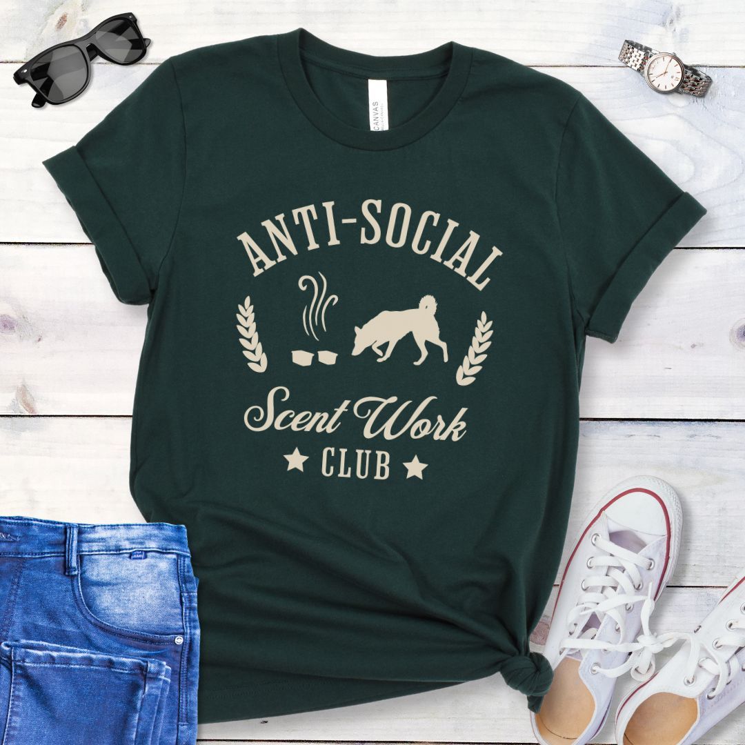 Dark green K9 nosework t-shirt with 'Anti-Social Scent Work Club' text and graphic of dog sniffing scentwork hides, on a wooden surface with jeans and shoes.