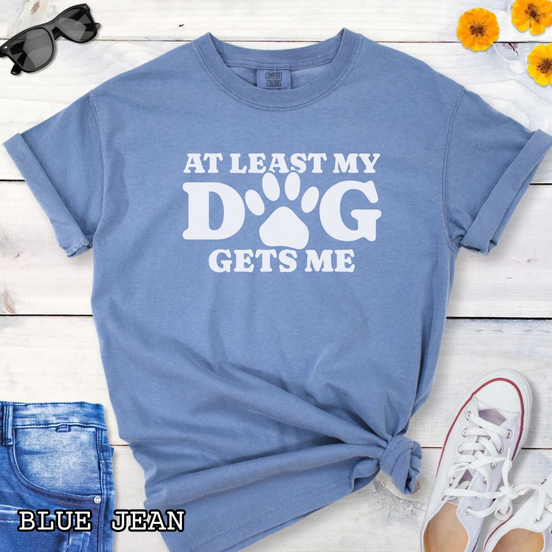 Blue Comfort Colors t-shirt for dog lovers with humorous text 'At least my dog gets me' in bold lettering with a paw print, shown on a wooden surface with sunglasses, jeans, and sneakers.