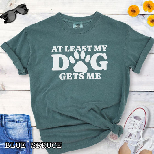 Green Comfort Colors t-shirt for dog lovers with humorous text 'At least my dog gets me' in bold lettering with a paw print, shown on a wooden surface with sunglasses, jeans, and sneakers.