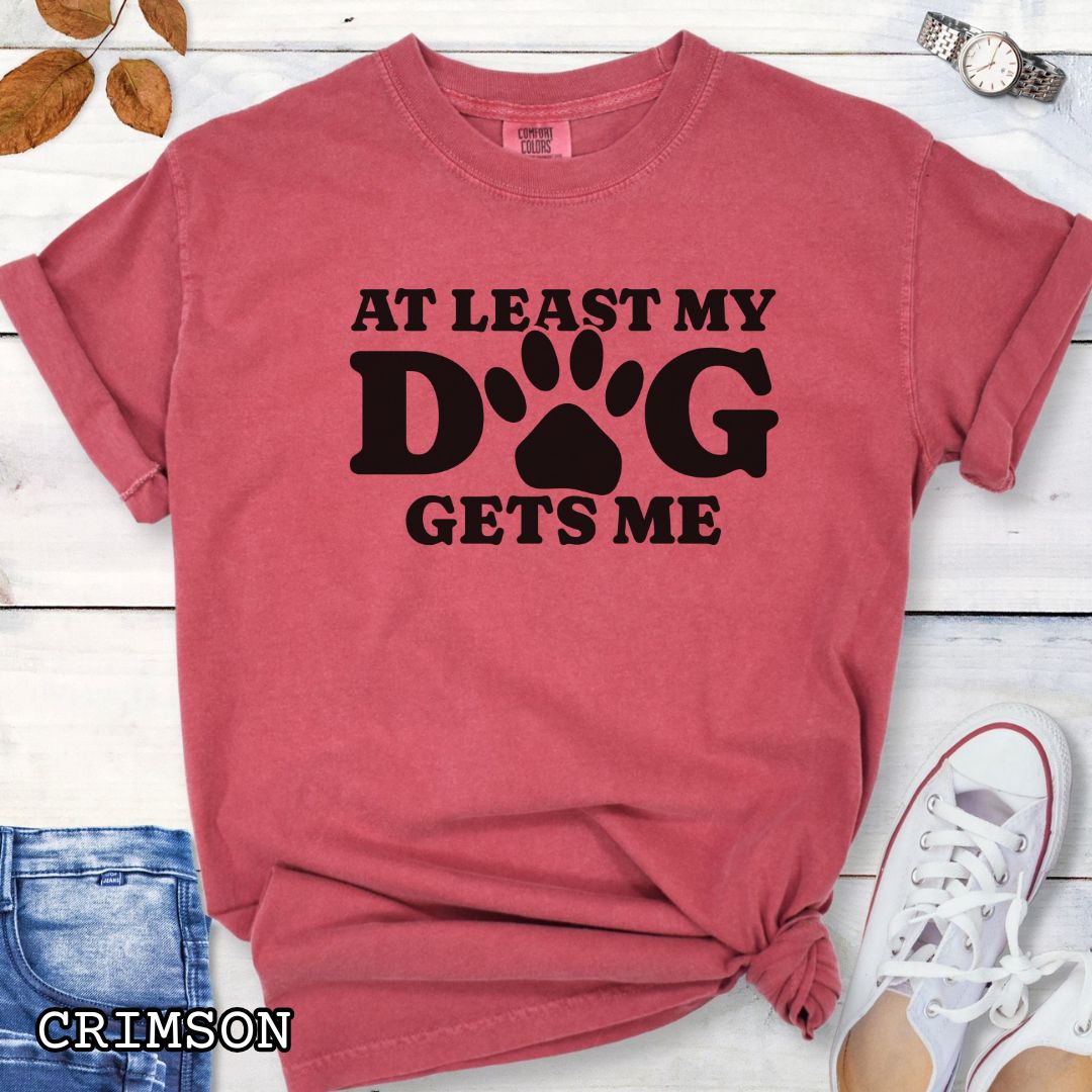 Red Comfort Colors t-shirt for dog lovers with humorous text 'At least my dog gets me' in bold lettering with a paw print, shown on a wooden surface with sunglasses, jeans, and sneakers.