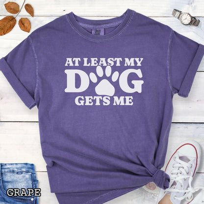 Purple Comfort Colors t-shirt for dog lovers with humorous text 'At least my dog gets me' in bold lettering with a paw print, shown on a wooden surface with sunglasses, jeans, and sneakers.