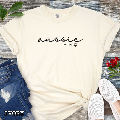 Beige Comfort Colors t-shirt for Australian Shepherd lovers with 'Aussie Mom' text design in feminine handwritten script, accompanied by a small paw print, shown on a wooden surface with sunglasses, jeans, and sneakers.