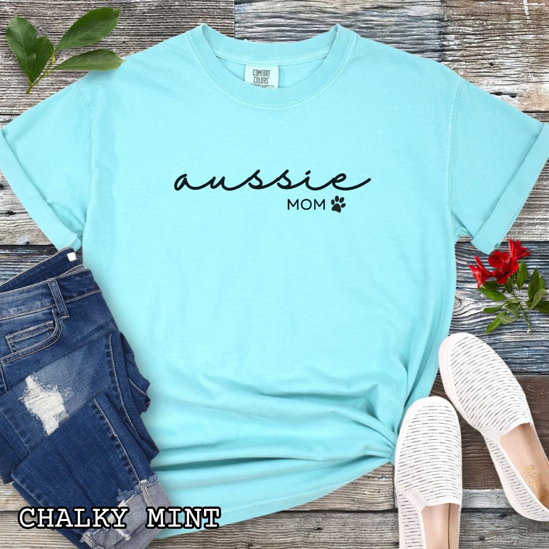 Mint Comfort Colors t-shirt for Australian Shepherd lovers with 'Aussie Mom' text design in feminine handwritten script, accompanied by a small paw print, shown on a wooden surface with sunglasses, jeans, and sneakers.