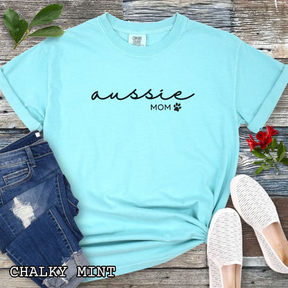 Mint Comfort Colors t-shirt for Australian Shepherd lovers with 'Aussie Mom' text design in feminine handwritten script, accompanied by a small paw print, shown on a wooden surface with sunglasses, jeans, and sneakers.