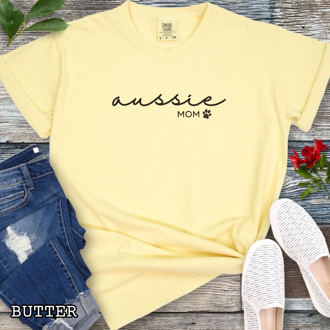 Yellow Comfort Colors t-shirt for Australian Shepherd lovers with 'Aussie Mom' text design in feminine handwritten script, accompanied by a small paw print, shown on a wooden surface with sunglasses, jeans, and sneakers.