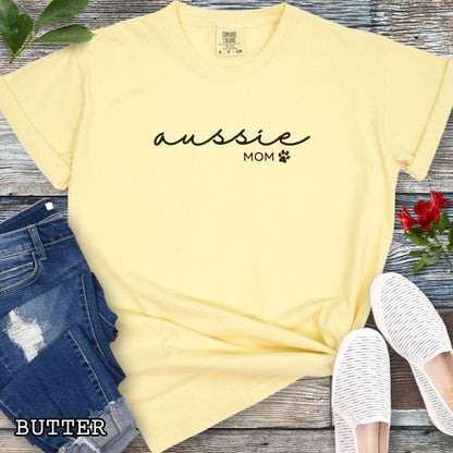 Yellow Comfort Colors t-shirt for Australian Shepherd lovers with 'Aussie Mom' text design in feminine handwritten script, accompanied by a small paw print, shown on a wooden surface with sunglasses, jeans, and sneakers.