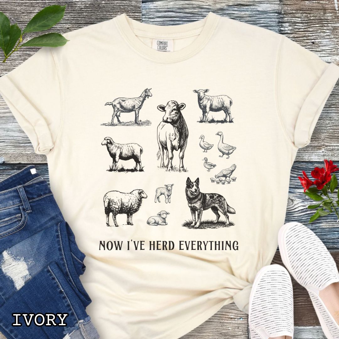 Beige Comfort Colors t-shirt for Australian Cattle Dog lovers with vintage  illustrations of farm animals and a Heeler dog, with the humorous text 'Now I've Herd Everything', shown on a wooden surface with denim jeans and flowers.