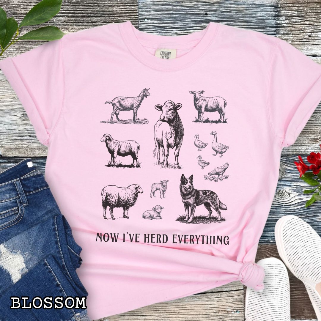 Pink Comfort Colors t-shirt for Australian Cattle Dog lovers with vintage  illustrations of farm animals and a Heeler dog, with the humorous text 'Now I've Herd Everything', shown on a wooden surface with denim jeans and flowers.
