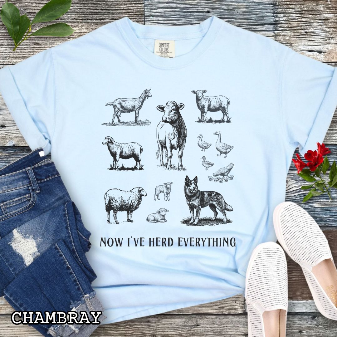 Light blue Comfort Colors t-shirt for Australian Cattle Dog lovers with vintage  illustrations of farm animals and a Heeler dog, with the humorous text 'Now I've Herd Everything', shown on a wooden surface with denim jeans and flowers.