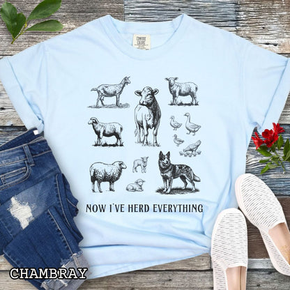 Light blue Comfort Colors t-shirt for Australian Cattle Dog lovers with vintage  illustrations of farm animals and a Heeler dog, with the humorous text 'Now I've Herd Everything', shown on a wooden surface with denim jeans and flowers.