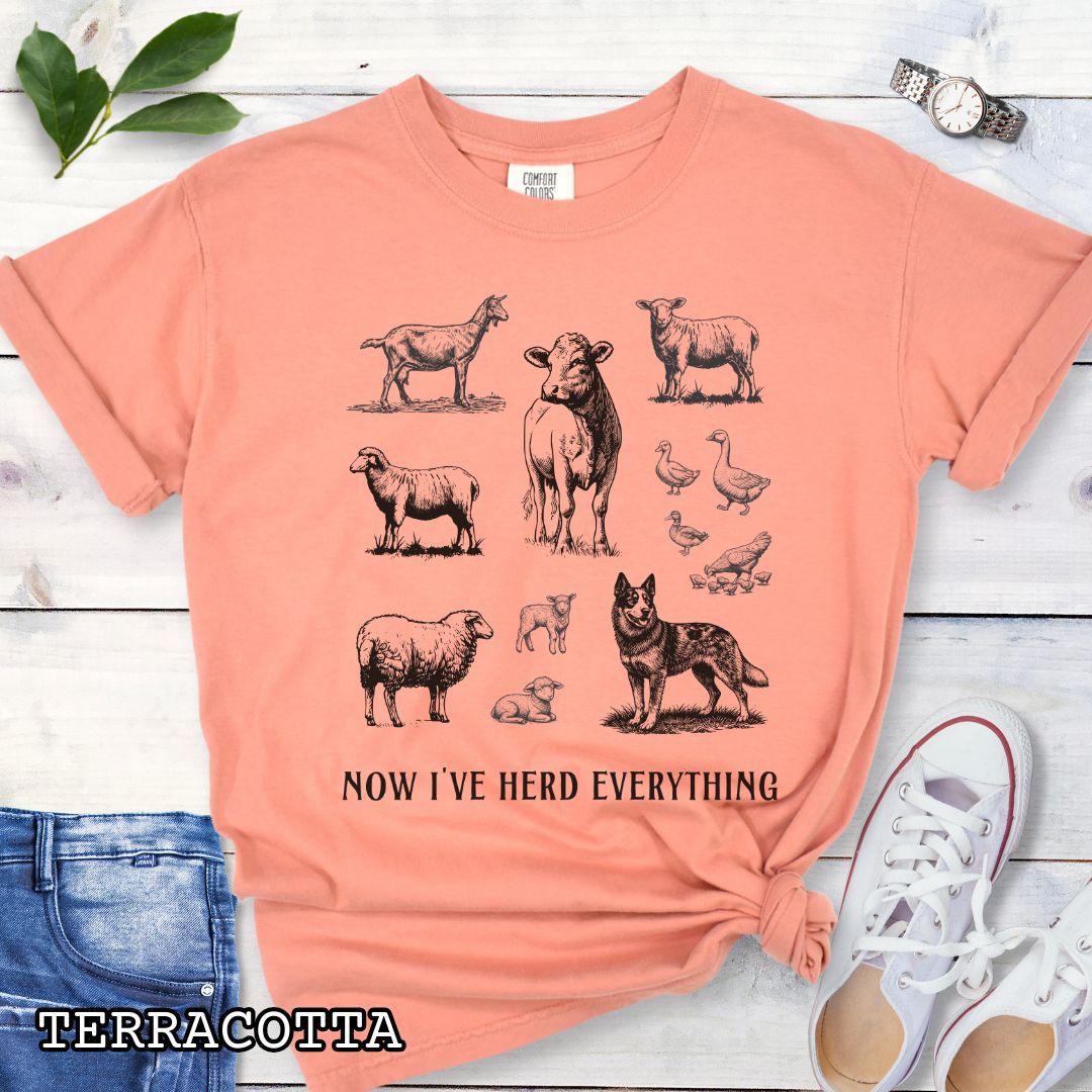 Orange Comfort Colors t-shirt for Australian Cattle Dog lovers with vintage  illustrations of farm animals and a Heeler dog, with the humorous text 'Now I've Herd Everything', shown on a wooden surface with denim jeans and flowers.