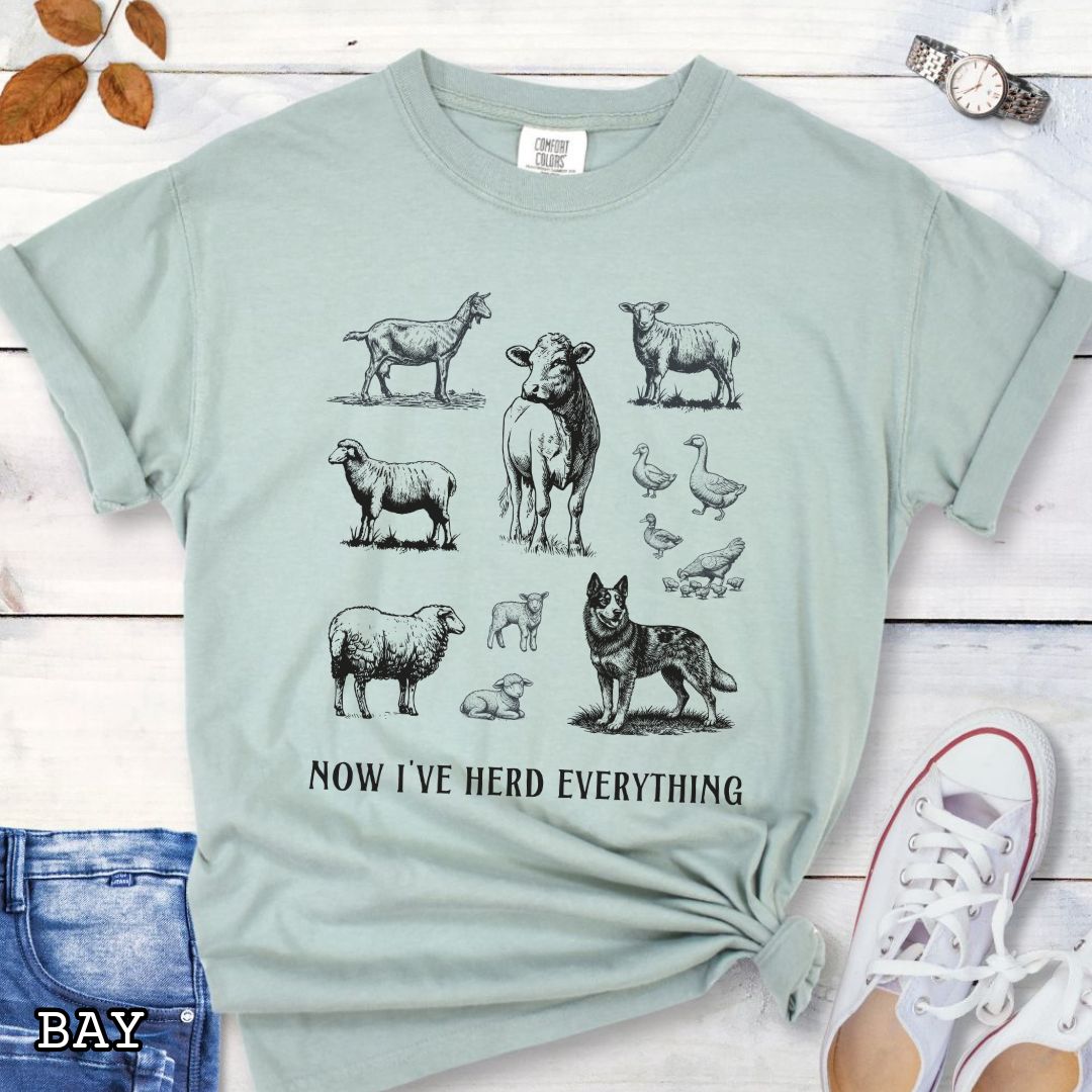 Light green Comfort Colors t-shirt for Australian Cattle Dog lovers with vintage  illustrations of farm animals and a Heeler dog, with the humorous text 'Now I've Herd Everything', shown on a wooden surface with denim jeans and flowers.