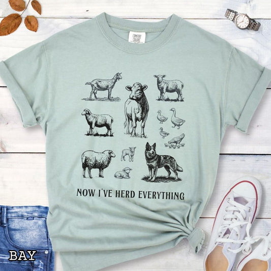 Light green Comfort Colors t-shirt for Australian Cattle Dog lovers with vintage  illustrations of farm animals and a Heeler dog, with the humorous text 'Now I've Herd Everything', shown on a wooden surface with denim jeans and flowers.