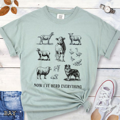 Light green Comfort Colors t-shirt for Australian Shepherd lovers with vintage farm animal illustrations and an Australian Shepherd dog, with the humorous text 'Now I've Herd Everything', shown on a wooden surface with denim jeans and flowers.