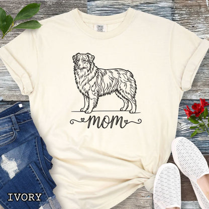 Beige Comfort Colors t-shirt with a line art Australian Shepherd dog illustration and 'mom' text in feminine script beneath, shown on a wooden surface with denim jeans and a flower.