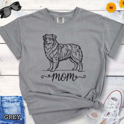 Grey Comfort Colors t-shirt with a line art Australian Shepherd dog illustration and 'mom' text in feminine script beneath, shown on a wooden surface with denim jeans and a flower.