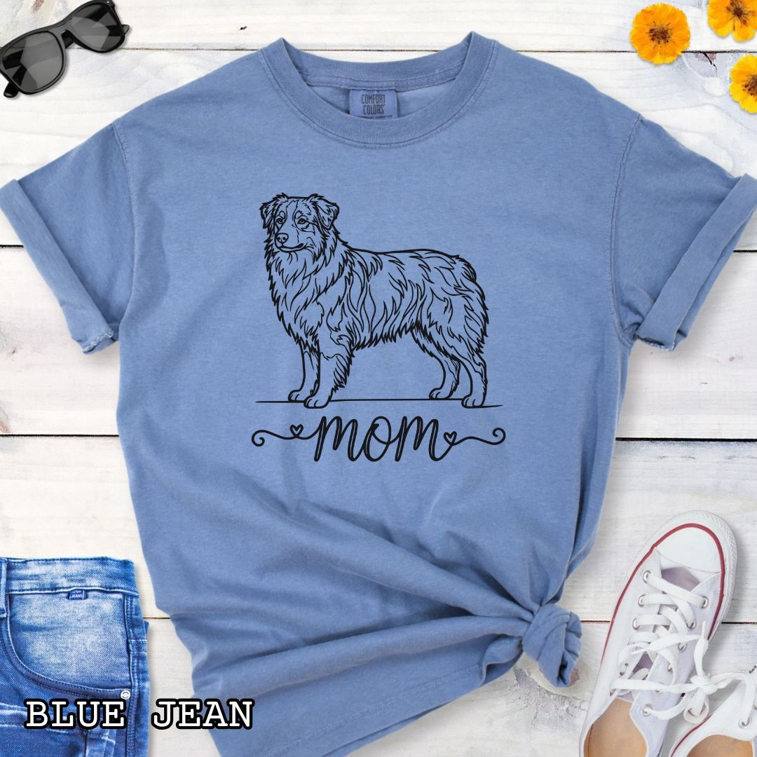 Blue Comfort Colors t-shirt with a line art Australian Shepherd dog illustration and 'mom' text in feminine script beneath, shown on a wooden surface with denim jeans and a flower.