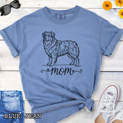 Blue Comfort Colors t-shirt with a line art Australian Shepherd dog illustration and 'mom' text in feminine script beneath, shown on a wooden surface with denim jeans and a flower.