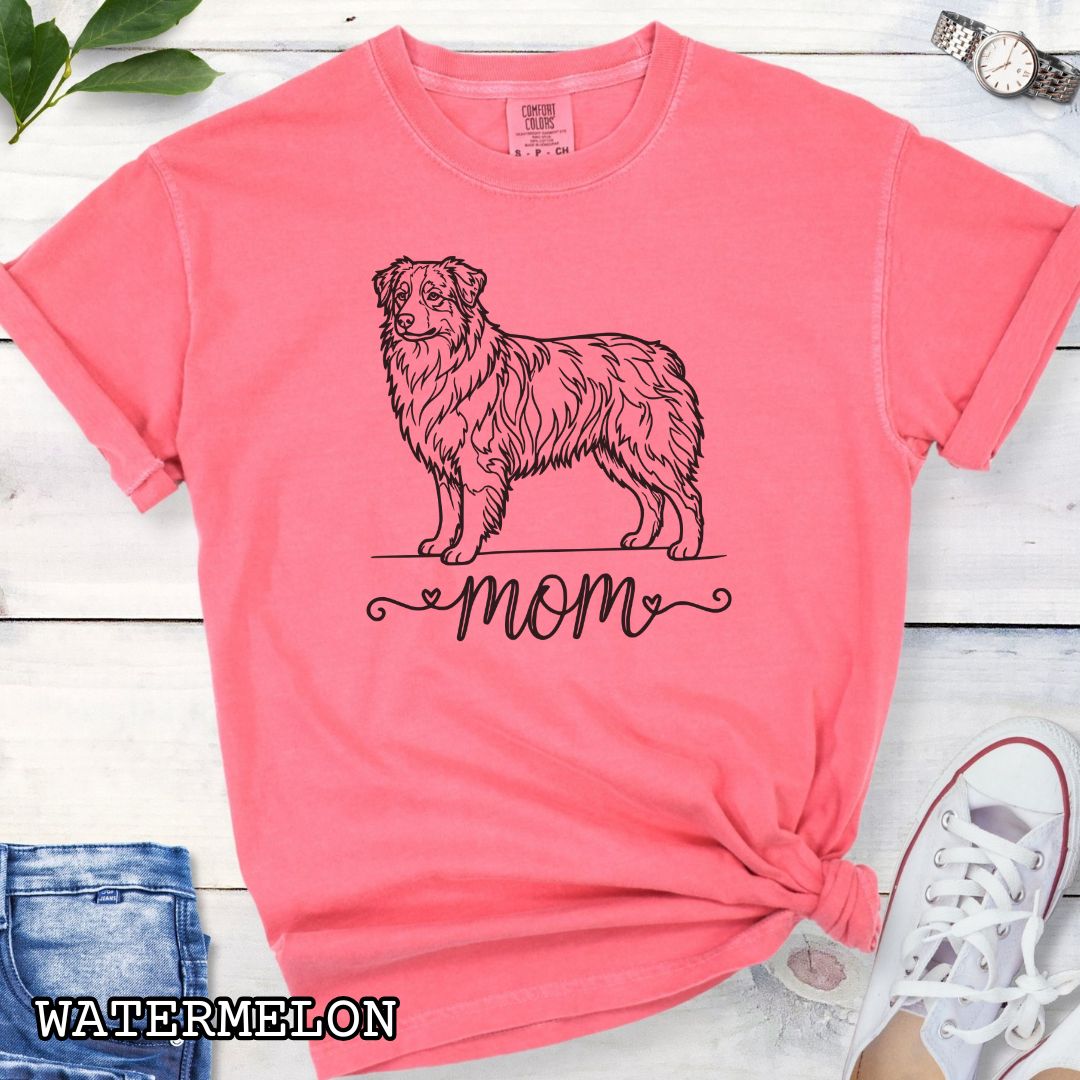 Pink Comfort Colors t-shirt with a line art Australian Shepherd dog illustration and 'mom' text in feminine script beneath, shown on a wooden surface with denim jeans and a flower.